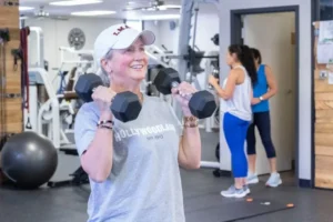 Woman lifting dumbbells in a gym with others exercising in the background.